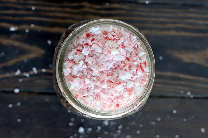 Overhead shot of a glass jar filled with crushed candy canes, on a dark brown wood surface with scattered white crumbs.