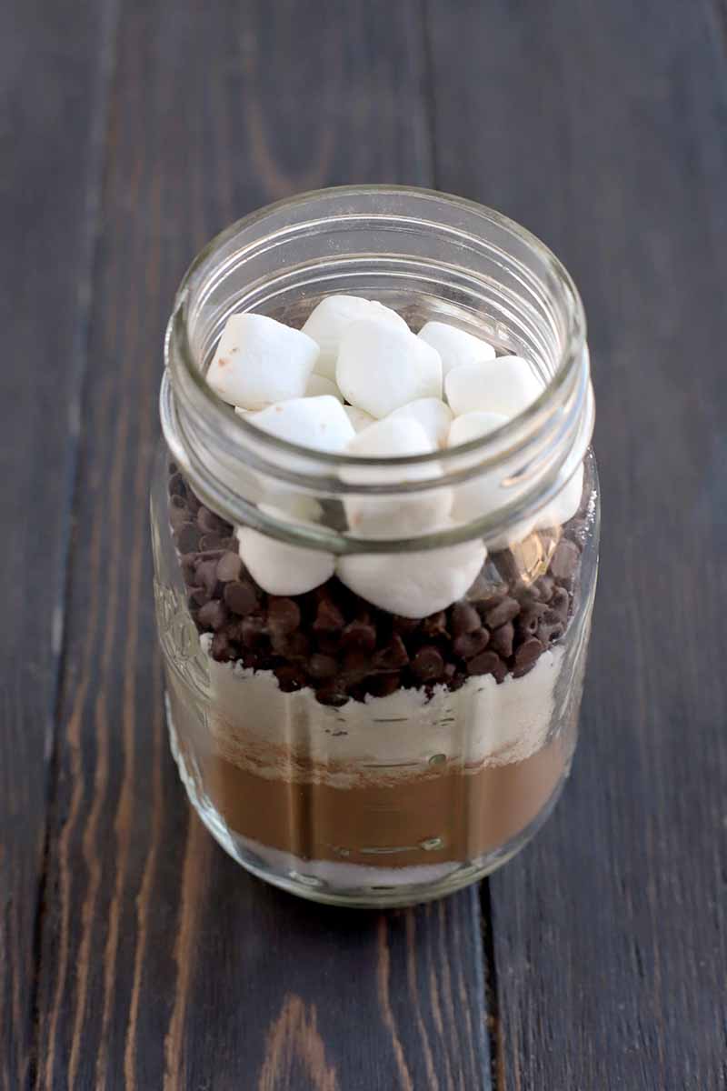 A glass jar is filled with layers of sugar, cocoa powder, powdered milk, mini chocolate chips, and mini marshmallows, on a dark brown wood table.