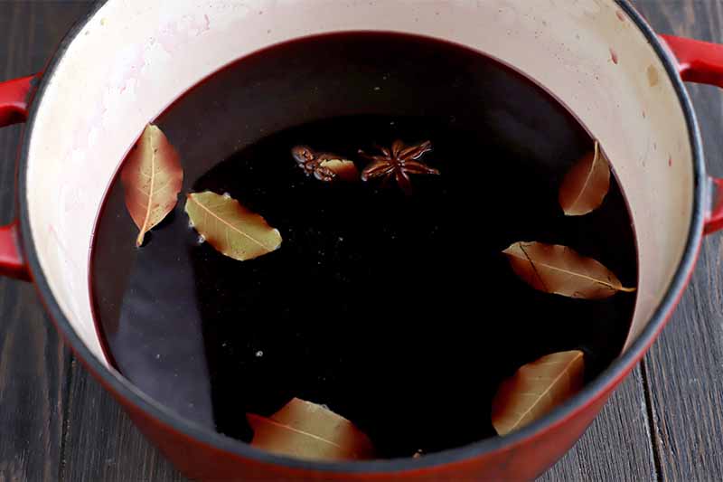 Dried bay leaves and star anise are floating in red wine in a red enameled pot with a cream-colored enameled interior, on a dark brown wood surface.