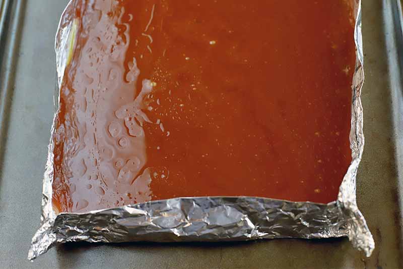 A homemade vegan toffee mixture is spread in a single, glassy layer in a square container made of aluminum foil, on a metal cookie sheet.