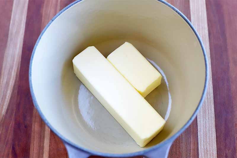 One and a half stick of butter in the bottom of a blue and white enameled saucepan, on a striped beige and brown wood background.