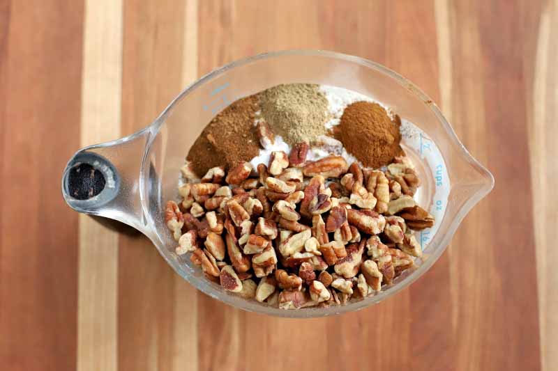 Overhead shot of chopped pecans and ground spices in a plastic pitcher-style measuring cup on a striped beige and brown wood background.