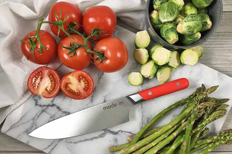 Horizontal image of a marble cutting board with sliced tomatoes, Brussels sprouts, and asparagus with a knife with a red handle.