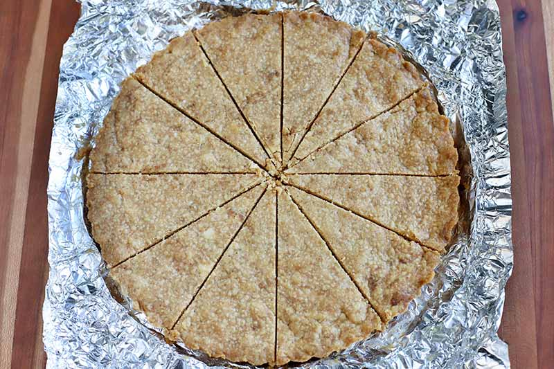 Overhead shot of a circular piece of homemade shortbread on top of a piece of foil, sliced into wedges, on a brown wood surface.