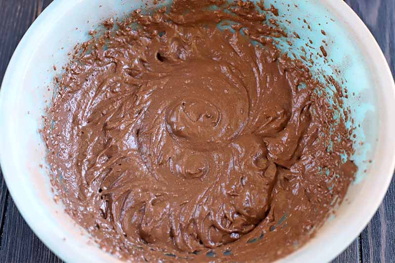 Overhead closely cropped shot of chocolate batter in a pale blue glass bowl, on a dark brown wood background.