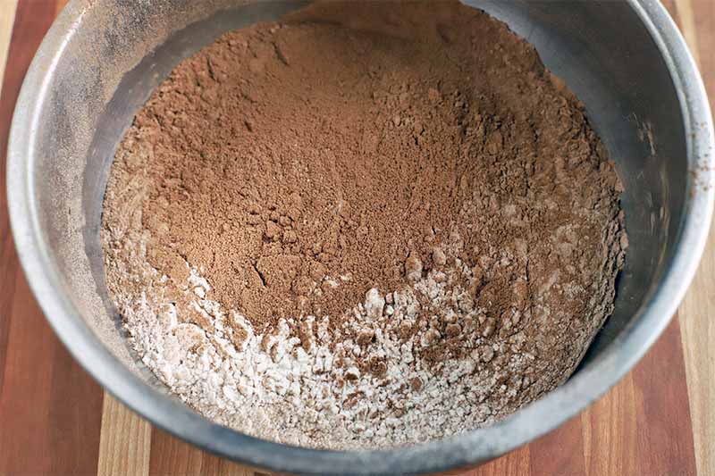 Sifted cocoa powder and other dry baking ingredients in a stainless steel bowl on a striped wood surface.