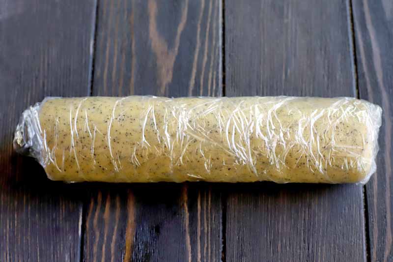 A log-shaped piece of cookie dough is wrapped tightly in plastic, and is resting on a dark brown wood table.
