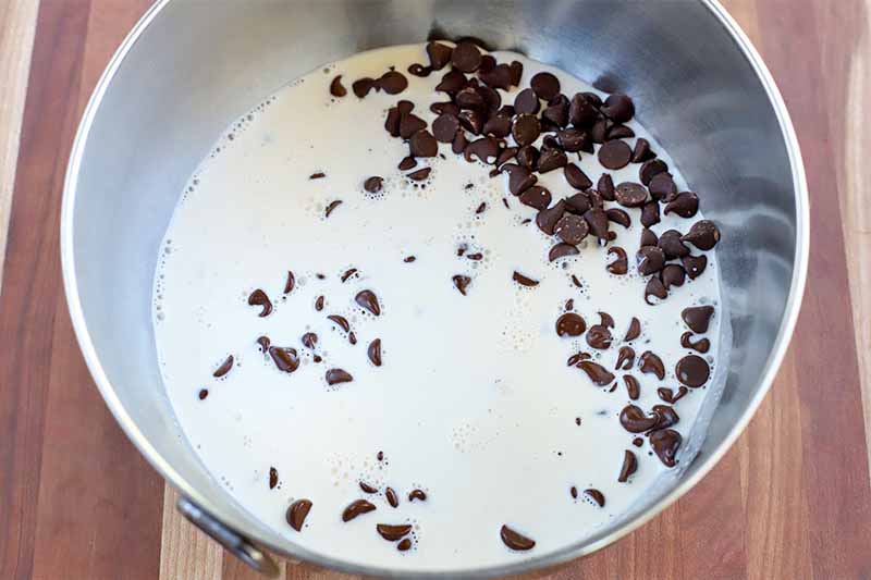 Milk and chocolate chips in a stainless steel mixing bowl, on a striped brown and beige wood surface.