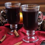 Two glass mugs of mulled red wine, on a red cloth surface with whole cinnamon sticks, citrus fruits, and decorative pine cones, with a brown wood background.