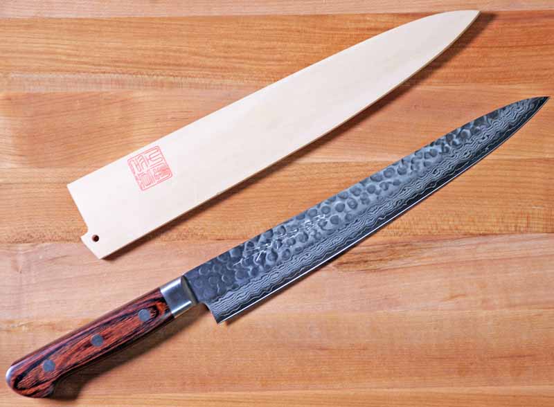 Top-down view of the Norisada 9.5-Inch Hammered Damascus Sujihiki with its wooden saya scabbard on a maple butcher block surface.