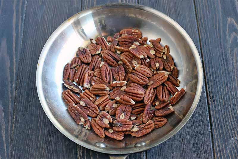Raw, unsalted pecan halves in a stainless steel frying pan, on a dark brown wood surface.
