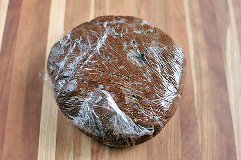 A flatterned disc of chocolate dough tightly wrapped in plastic, on a wood countertop.