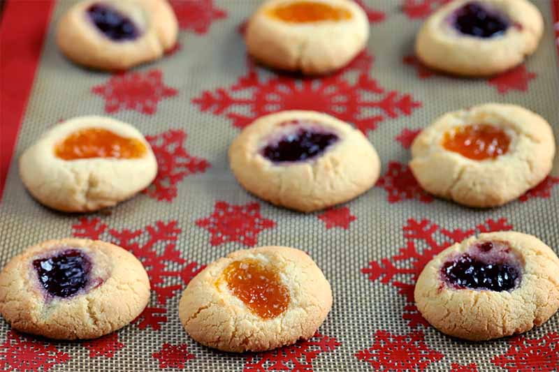 Oblique shot of thumbprint cookies filled with orange and purple-colored jam arranged on a red snowflake-patterned silicone baking sheet liner.