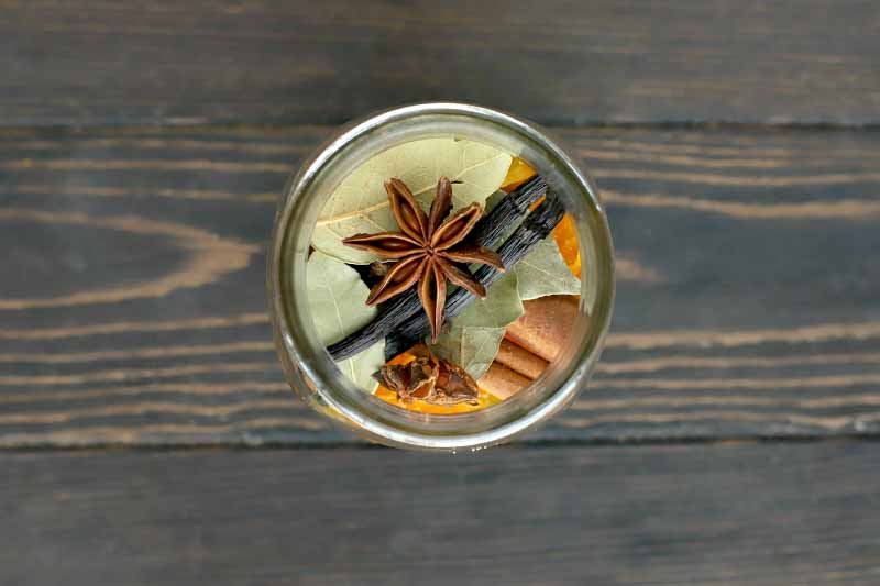 Overhead shot of a glass jar filled iwth whole bay leaves, star anise, citrus peel, cinnamon sticks, and vanilla beans, on a dark brown wood table.