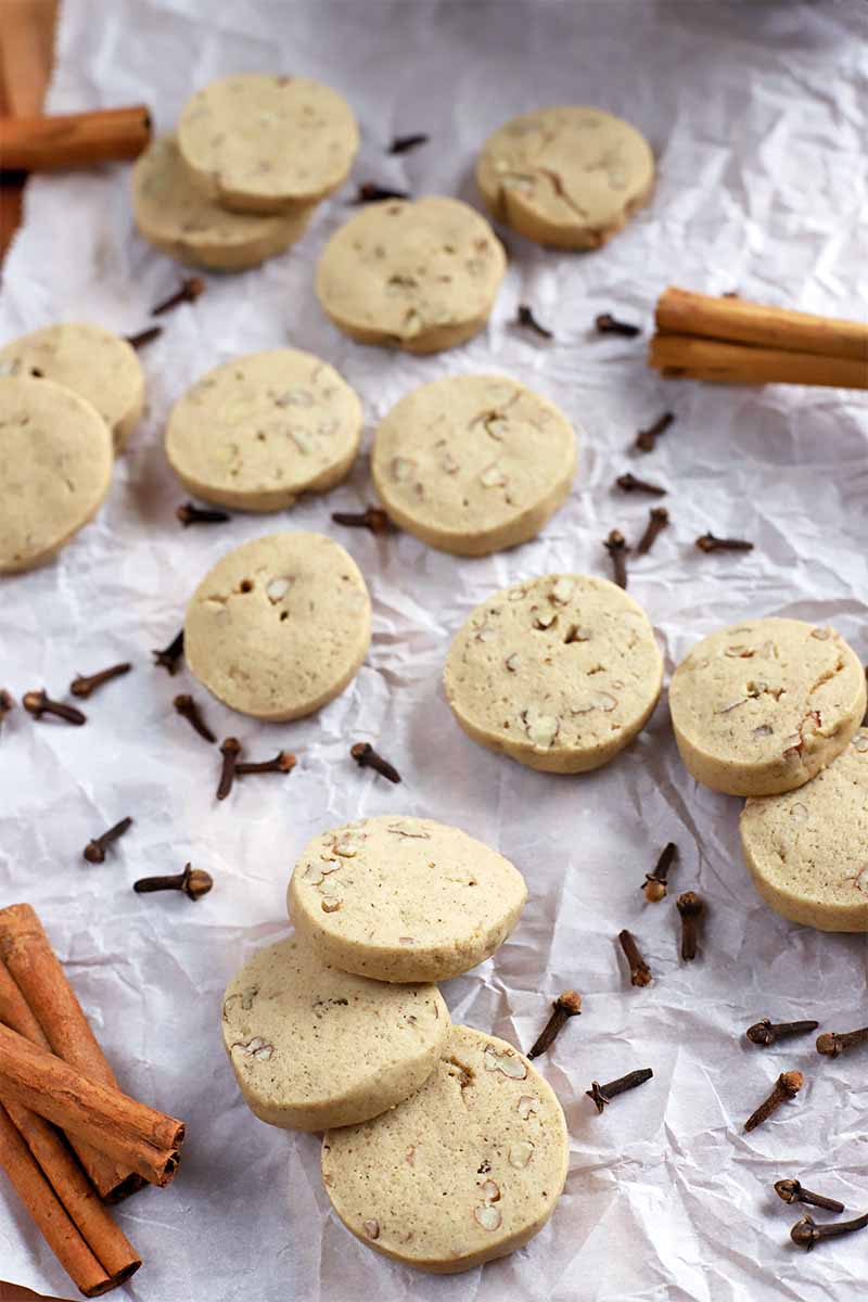 Overhead oblique vertical shot of scattered round cookies with whole cloves and cinnamon sticks on a piece of parchment paper.