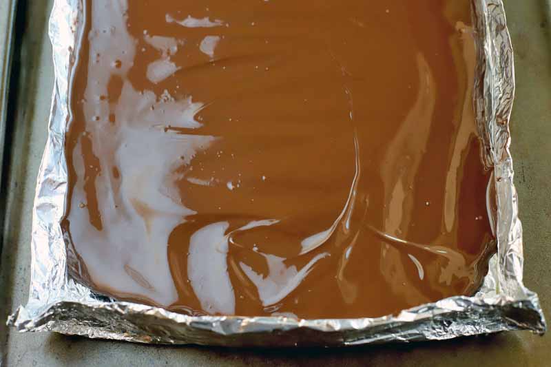 Melted chocolate is spread in an even layer in a square aluminum foil shell, on top of a metal sheet pan.