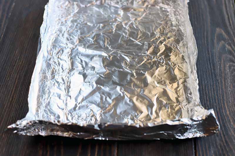 Aluminum foil is folded to make a square tray with sizes approximately 1 inch tall, on a dark brown wood table.