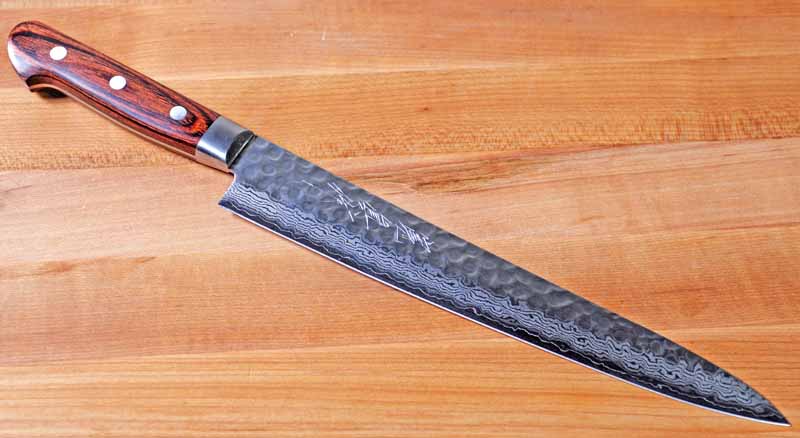 The Norisada 9.5-Inch Hammered Damascus Slicing Knife without its saya scabbard on a wooden cutting board. 