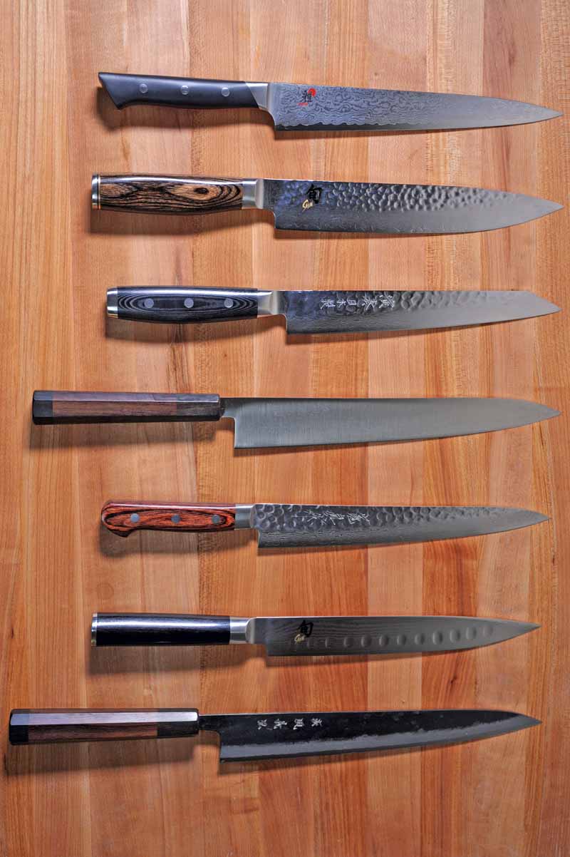 Vertical image of seven Japanese sujihiki knives arranged in a row on a wooden cutting board.