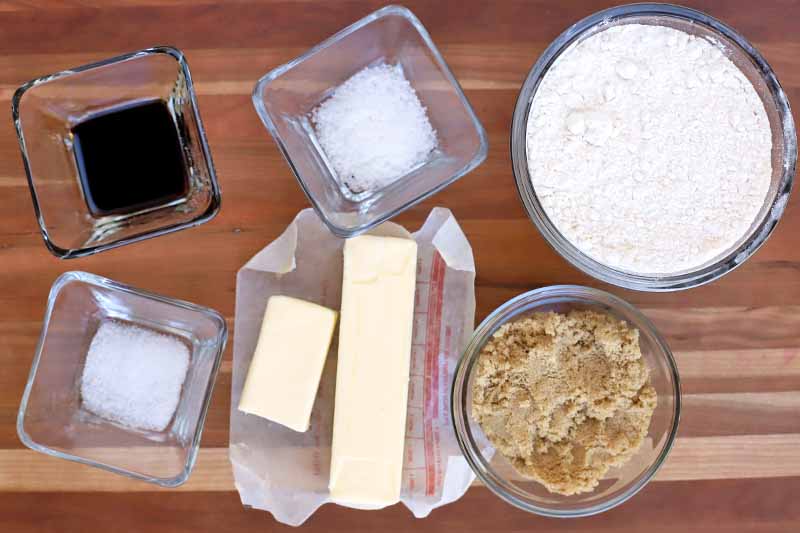 Overhead shot of three square and two round glass bowls of vanilla, salt, sugar, flour, and brown sugar, with butter on a waxed paper wrapper, on a striped brown wood surface.