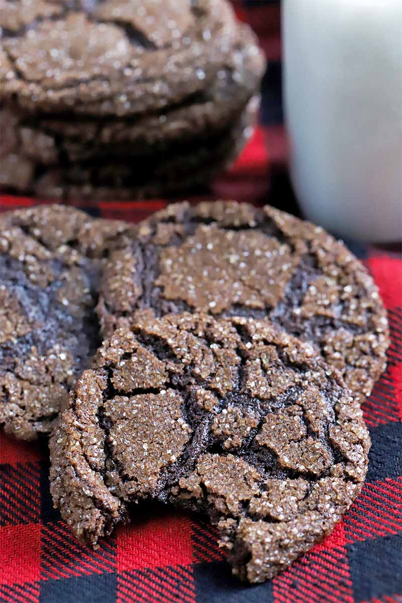 Three dark chocolate cookies are in the foreground with a short stack beside a glass of milk in the background, with a bite taken out of the one closest to the viewer, on a red and black flannel cloth.