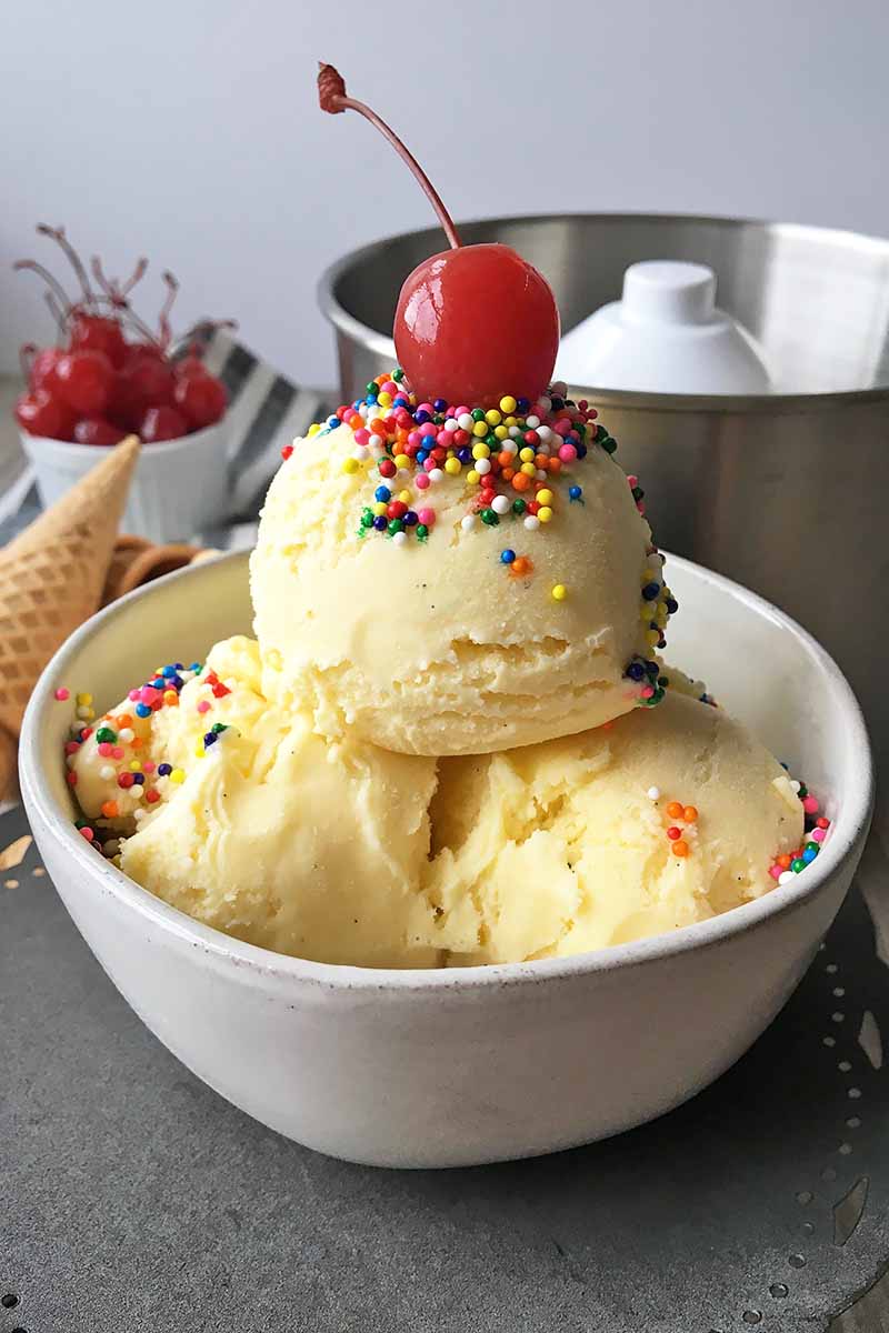 Vertical image of a bowl of ice cream with sprinkles and a cherry in front of an ice cream maker and a bowl of cherries and cones.