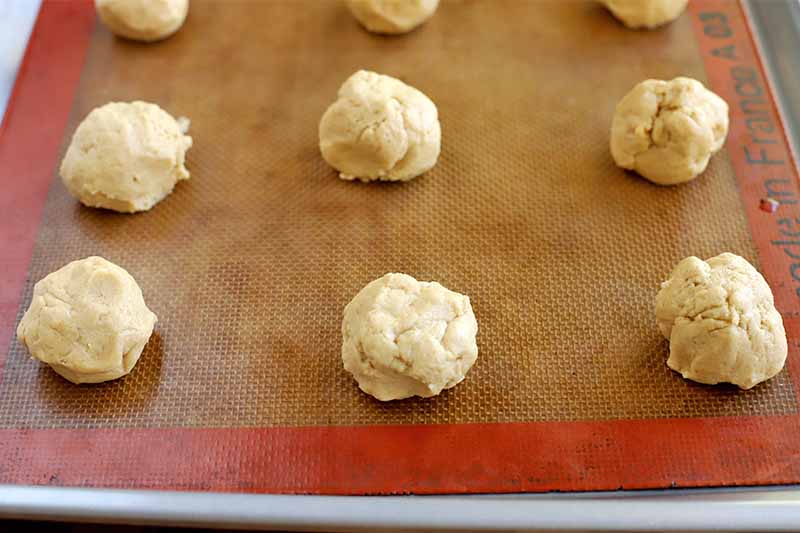Small balls of peanut butter cookie dough arranged in rows on an orange and beige Silpat silicone pan liner on a metal baking sheet pan.