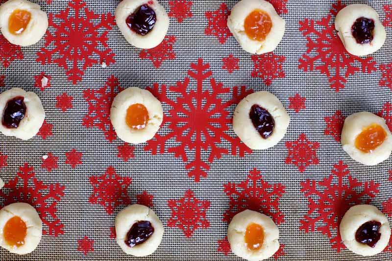 Over head shot of twelve almond thumbprint cookies filled with two types of fruit jam, arranged in three rows on a silicone pan liner decorated with a red snowflake pattern.