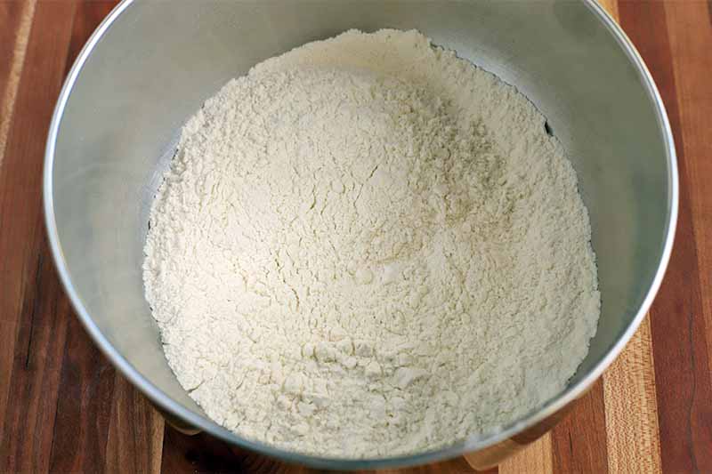 A dry flour mixture is at the bottom of a large stainless steel mixing bowl, on a striped wood surface.