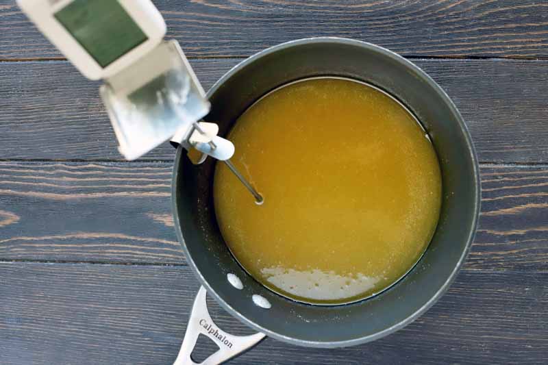 Overhead shot of a nonstick Calphalon saucepan filled with a margarine mixture, with a candy thermometer attached to the rim with a metal clip, on a dark brown wood surface.