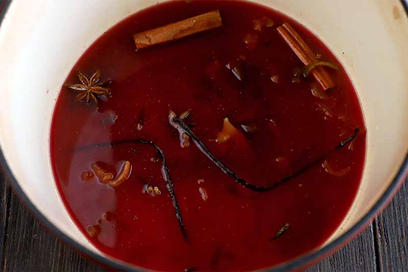 Top-down closely cropped image of an enameled stockpot with red wine, whole cinnamon sticks and star anise, vanilla beans, and orange peel at the bottom, on a dark brown wood surface.