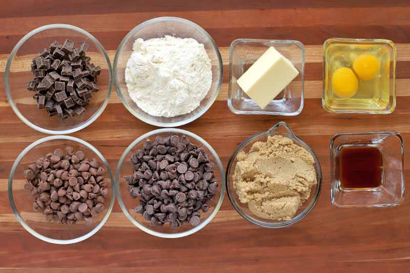 Overhead shot of square and round glass dishes of chocolate chips and chunks, flour, butter, eggs, brown sugar, and vanilla, on a striped brown wood table.