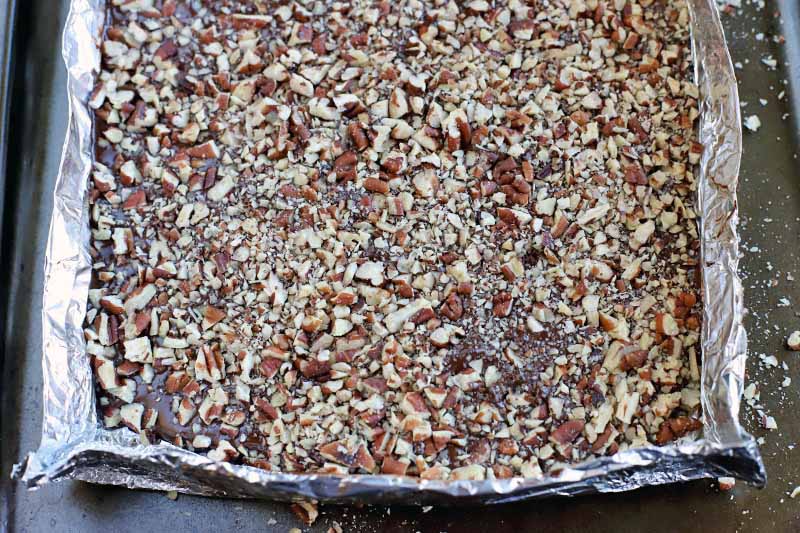 Aluminum foil is shaped into a square with sizes about 1 inch tall, with a layer of toasted pecans visible inside, on a metal baking pan.