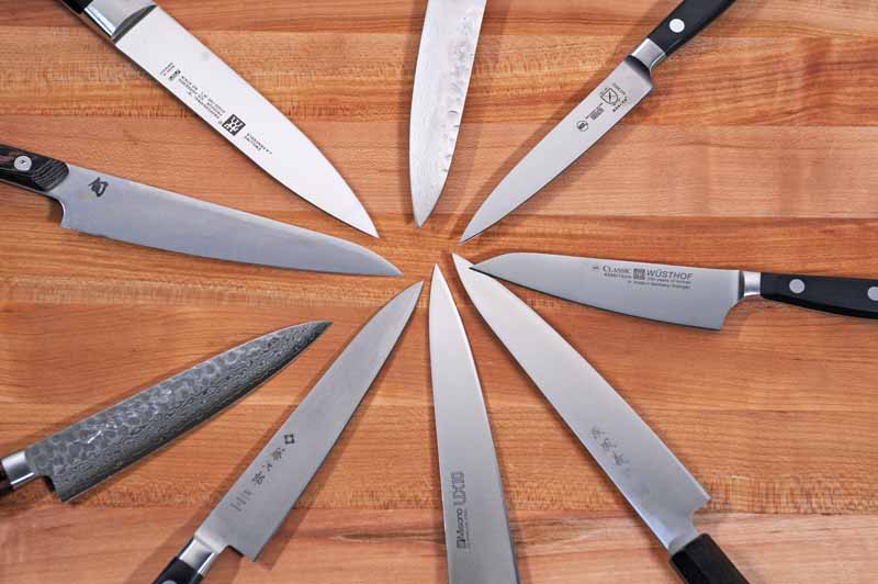 Top down view of 9 different kitchen utility and petty knives with their tips all pointed to one point and organized in a circle.