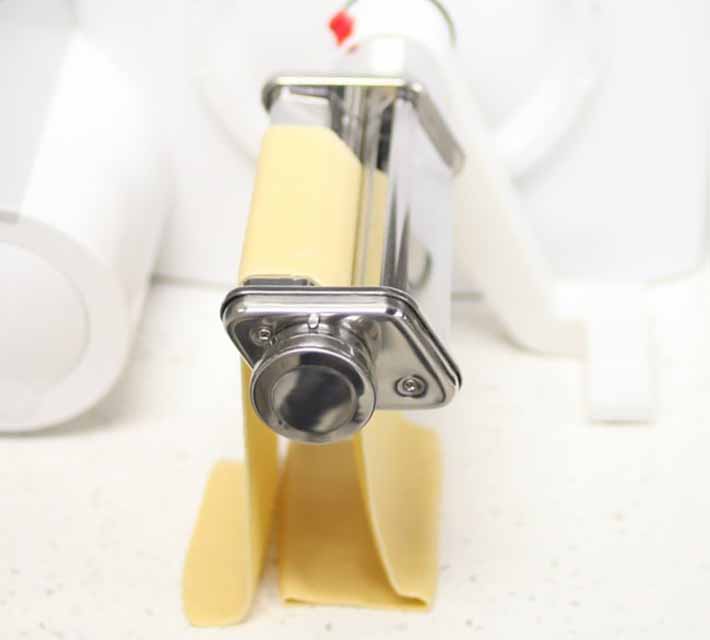 Square image of a pasta maker attachment on a mixing bowl base with a sheet of yellow pasta.