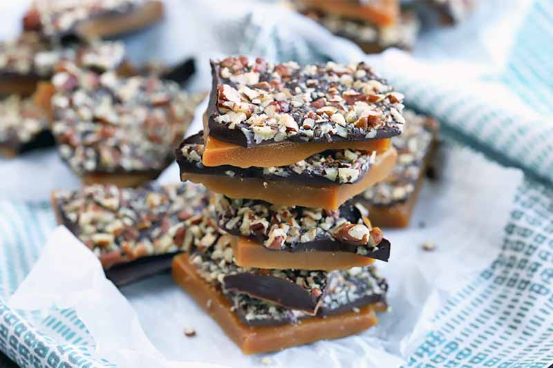 A stack of five pieces of homemade lactose-free toffee with dark chocolate and toasted pecans, on a piece of parchment paper on top of a folded and gathered light blue and white cloth, with more candy in soft focus in the background.