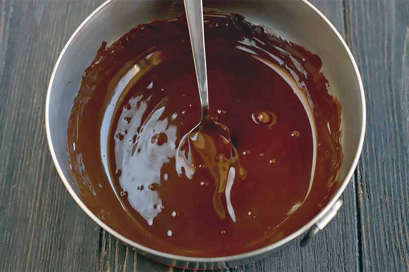 Top-down closely cropped shot of melted chocolate in a stainless steel mixing bowl with a spoon, on a dark brown wood surface.