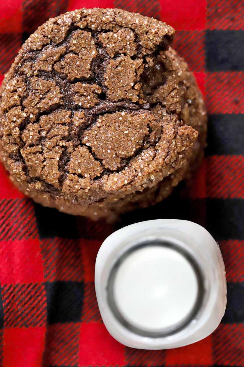 Overhead shot of a short stack of dark chocolate cookies with a bite taken out of the topmost one, with a glass bottle of milk, on a black and red plaid flannel cloth.