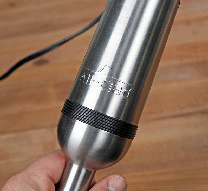 A closeup shot of the stainless steel body and motor assembly of the All-Clad KZ750D Immersion Blender, with a maple wooden butcher block background.