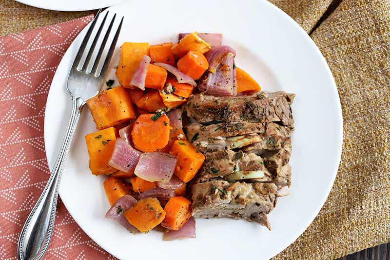 A plate of pork, sweet poato, carrots, and onions, with a fork resting on the rim, on top of tan burlap and a pink patterned cloth napkin.