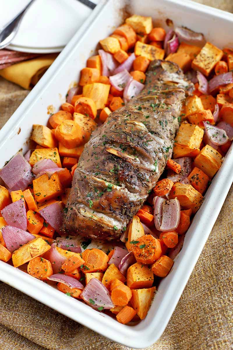 Roast pork tenderloin is at the center of a rectangular ceramic baking dish, surrounded by chopped roasted root vegetables, on a burlap surface with a yellow cloth napkin and white plates.