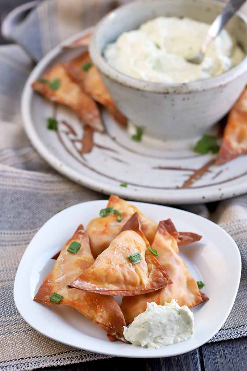 Vertical image of a white plate of homemade buffalo chicken wonton bites with blue cheese dip, with a platter and bowl of more of the same in the background, on a wood surface covered with a tan cloth.