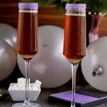 Sugar Plum Cocktails in two champagne flutes with dishes of sugar cubes and purple sanding sugar, silver balloons, and a large burgundy vase of evergreen branches in the background, against a brown backdrop.