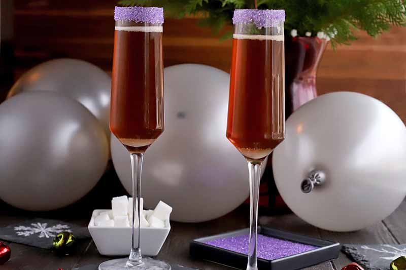 Sugar Plum Cocktails in two champagne flutes with dishes of white cubes and purple sanding sugar, silver balloons, and a large burgundy vase of evergreen branches in the background, against a brown backdrop.