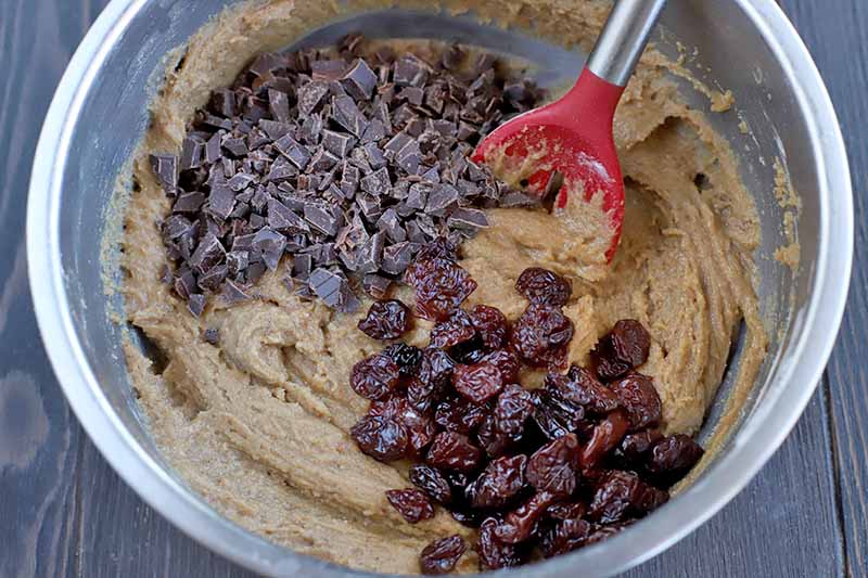 A red rubber slotted spoon with a metal handle is stirring a tan batter with two piles of dark chocolate chunks and dried cherries on top, in a stainless steel mixing bowl, on a dark brown wood surface.