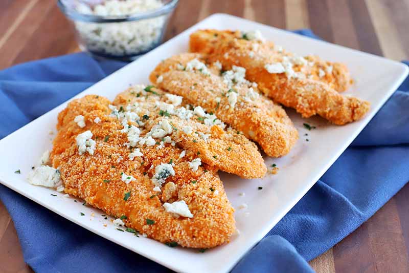 Four buffalo-style breaded chicken cutlets with cheese crumbles and fresh herbs on top, arranged on a square, white, ceramic serving platter on top of a blue cloth, on a striped brown wood surface with a small glass bowl of blue cheese towards the top left side of the frame.