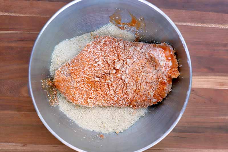 A piece of chicken breast has been dipped in a hot sauce and butter mixture and then dipped in a stainless steel bowl containing a mixture of bread crumbs and cheese, on a wood surface.
