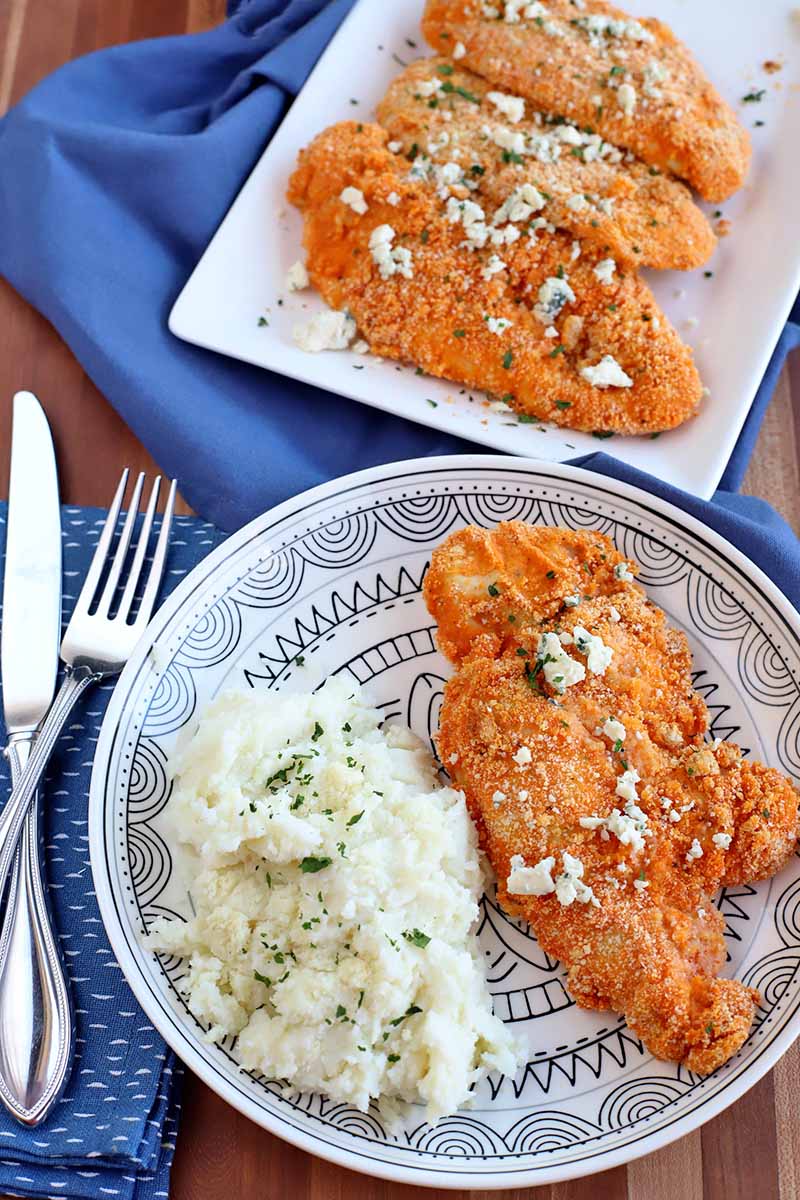 Vertical overhead shot of a plate and a serving platter of crispy buffalo chicken cutlets, with a serving of mashed potatoes topped with minced fresh herbs, with two blue cloth napkins and a knife and fork, on a brown wood surface.