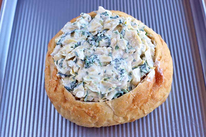 A boule loaf stuffed with spinach artichoke dip on a metal baking sheet with a ridged surface.