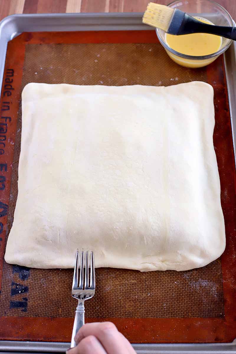 Overhead shot of a hand using a fork to crimp the edge of a square of filled puff pastry, on a Silpat silicone pan liner in a rimmed baking pan, with a small glass dish of egg wash with a beige and black silicone and plastic pastry brush resting on the rim, on a wood background.